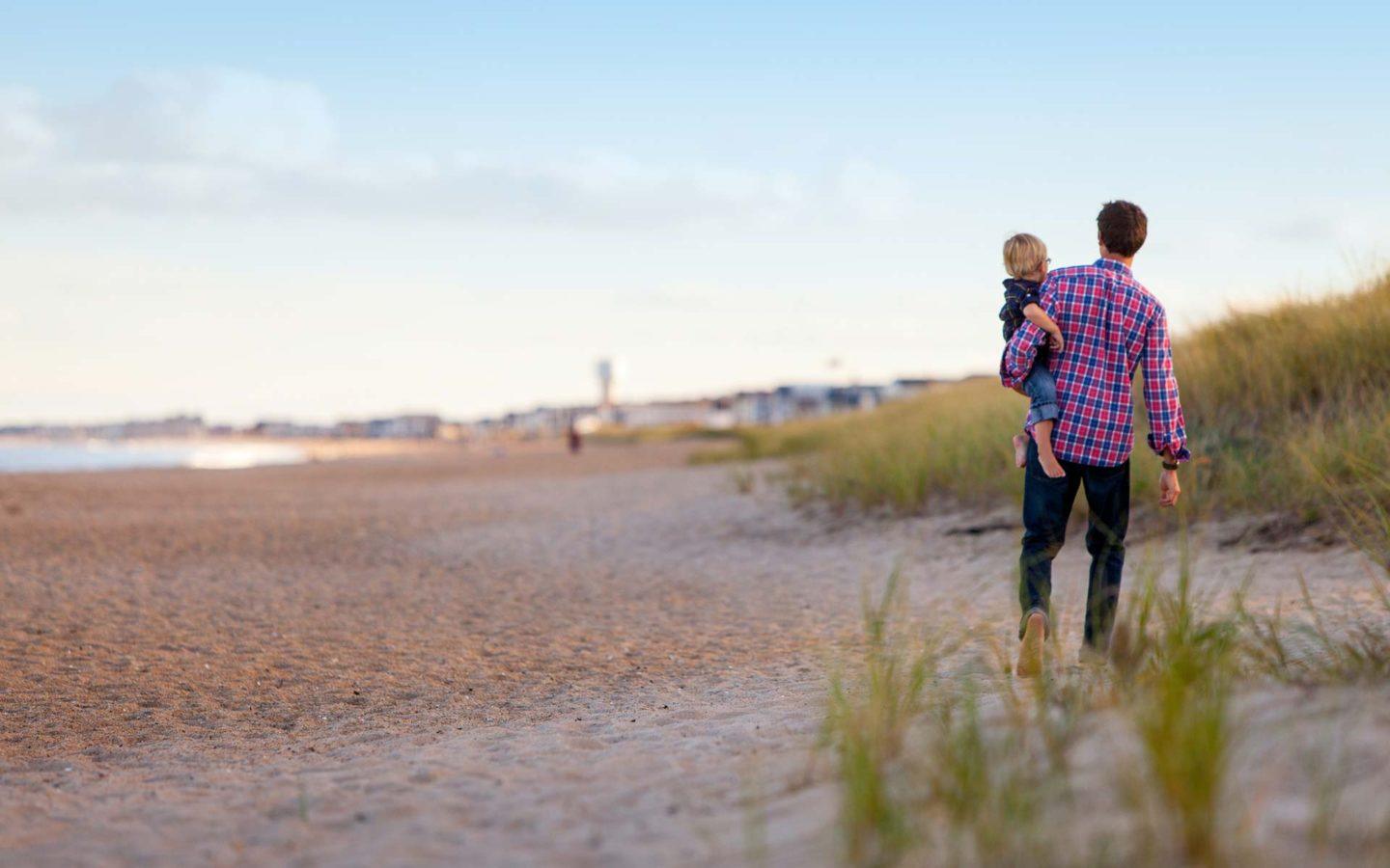 Man in flannel shirt holding his child while walking on a sandy beach