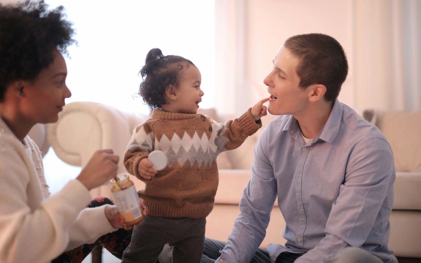 baby girl pointing finger at man's mouth, with women holding baby food